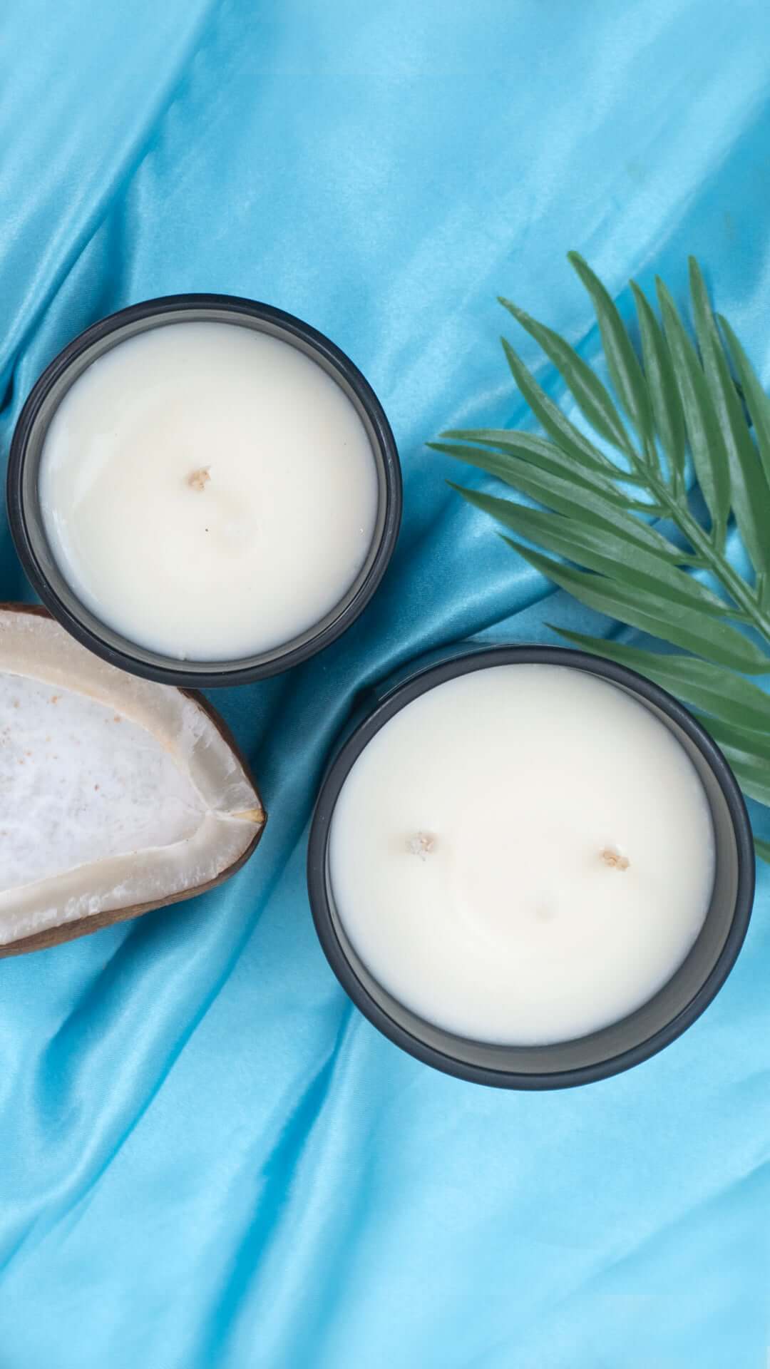 Seychelles scented candles in black containers on teal fabric with palm leaves and a coconut shell decor.